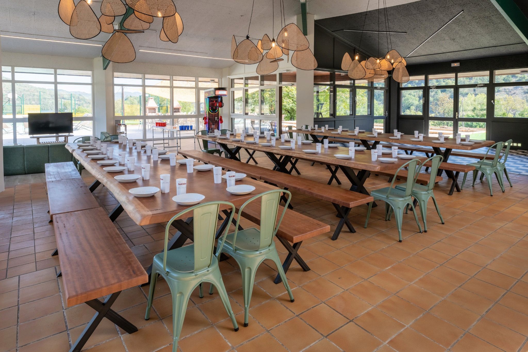 Un vaste espace dédié à la salle à manger, avec de longues tables en bois, des chaises vertes et des luminaires suspendus en rotin.