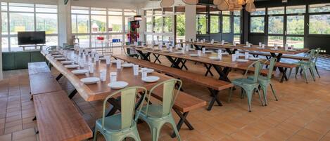 A spacious dining area with long wooden tables, green chairs, and hanging rattan light fixtures.