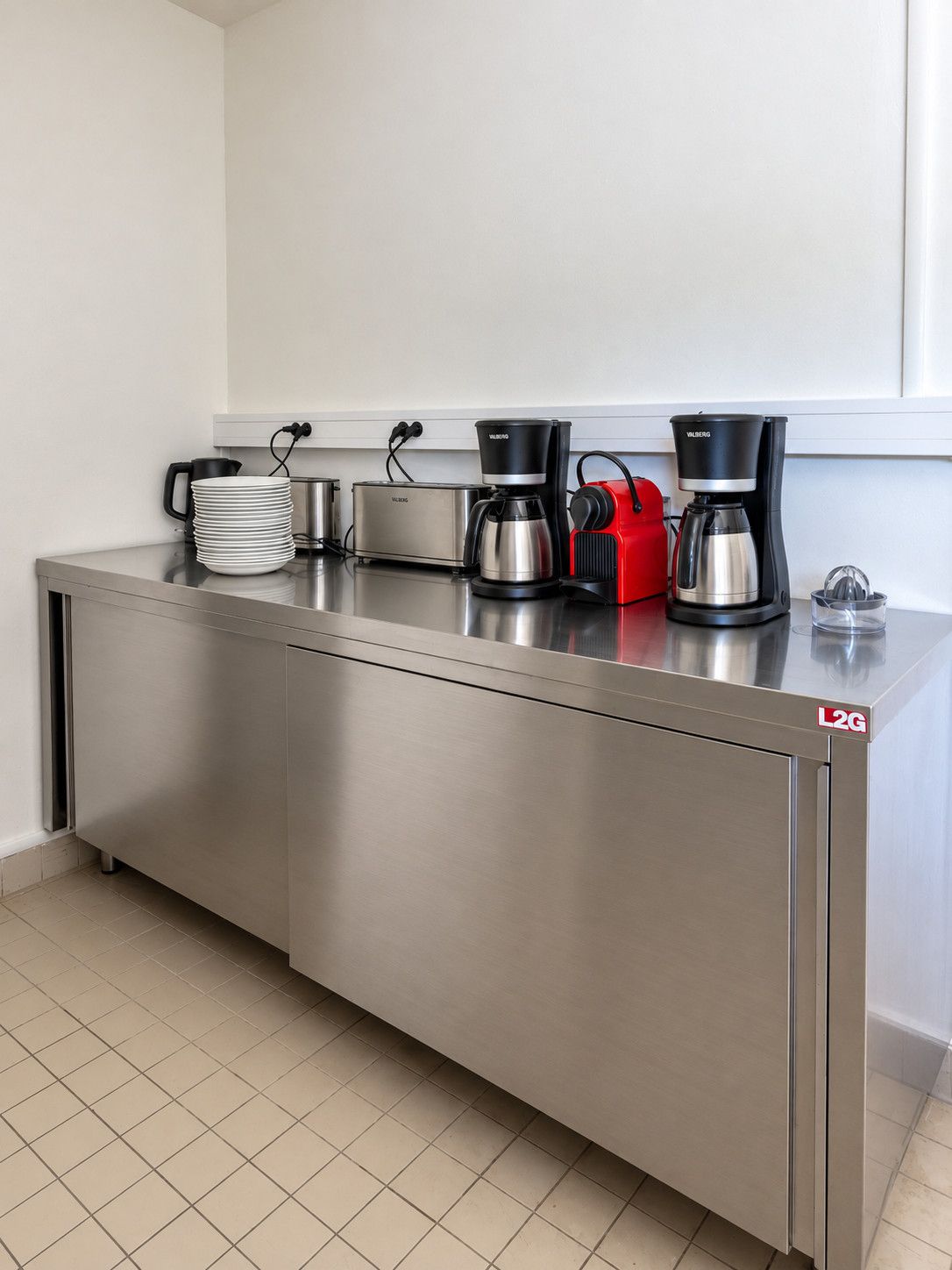 Une petite cuisine en acier inoxydable équipée d’une machine à café, d’un grille-pain et d’un appareil de cuisine rouge, probablement un cuiseur à riz ou un mijoteuse.