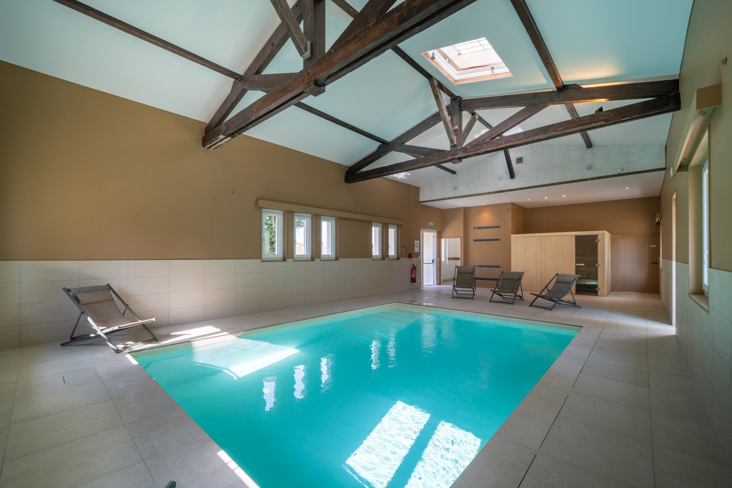 Une piscine intérieure dotée d’un haut plafond et de lucarnes, entourée de chaises longues et recouverte d’un sol carrelé.