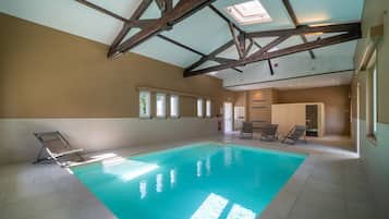 Une piscine intérieure dotée d’un haut plafond et de lucarnes, entourée de chaises longues et recouverte d’un sol carrelé.