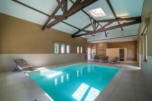 An indoor swimming pool with a high ceiling and skylights, surrounded by lounge chairs and a tiled floor.
