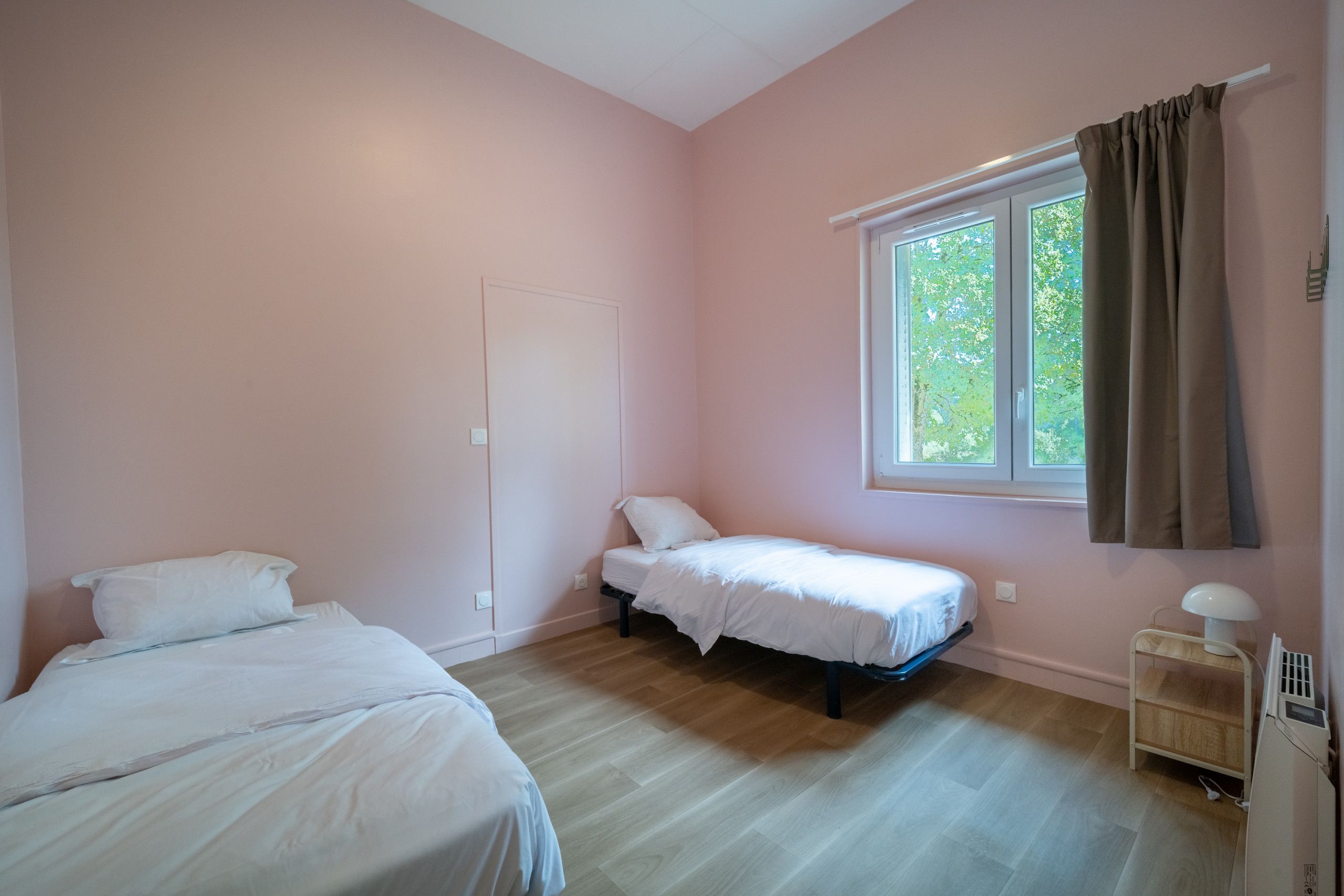 A bedroom with two beds, a window with curtains, and a bedside table with a lamp.