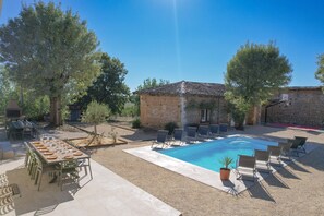 Area piscina esterna con tavolo e sedie, circondata da alberi e un edificio in pietra.