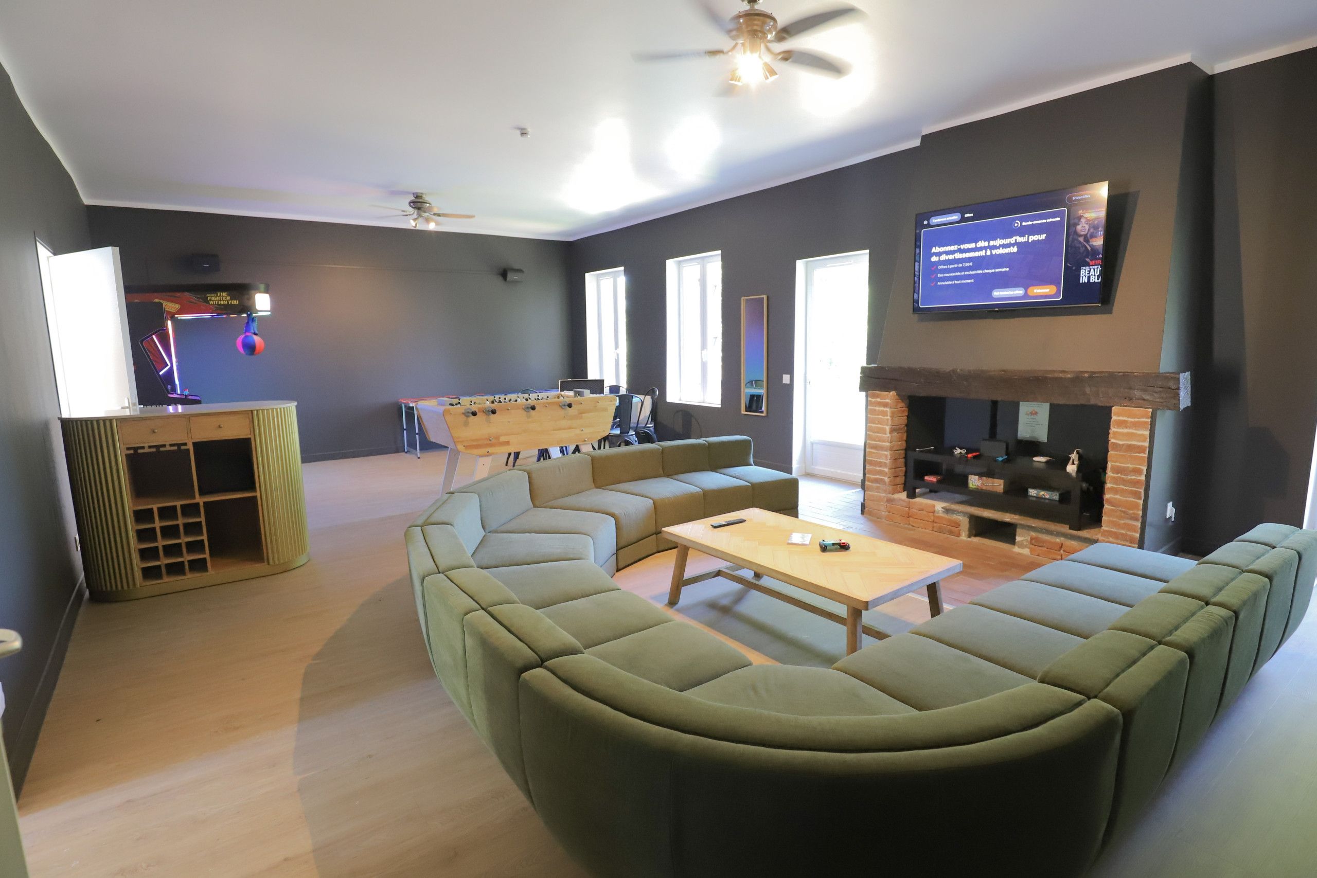 Een moderne woonkamer met een hoekbank, een salontafel, een tafelvoetbalspel, een flatscreen-tv aan de muur en een open haard met bakstenen.