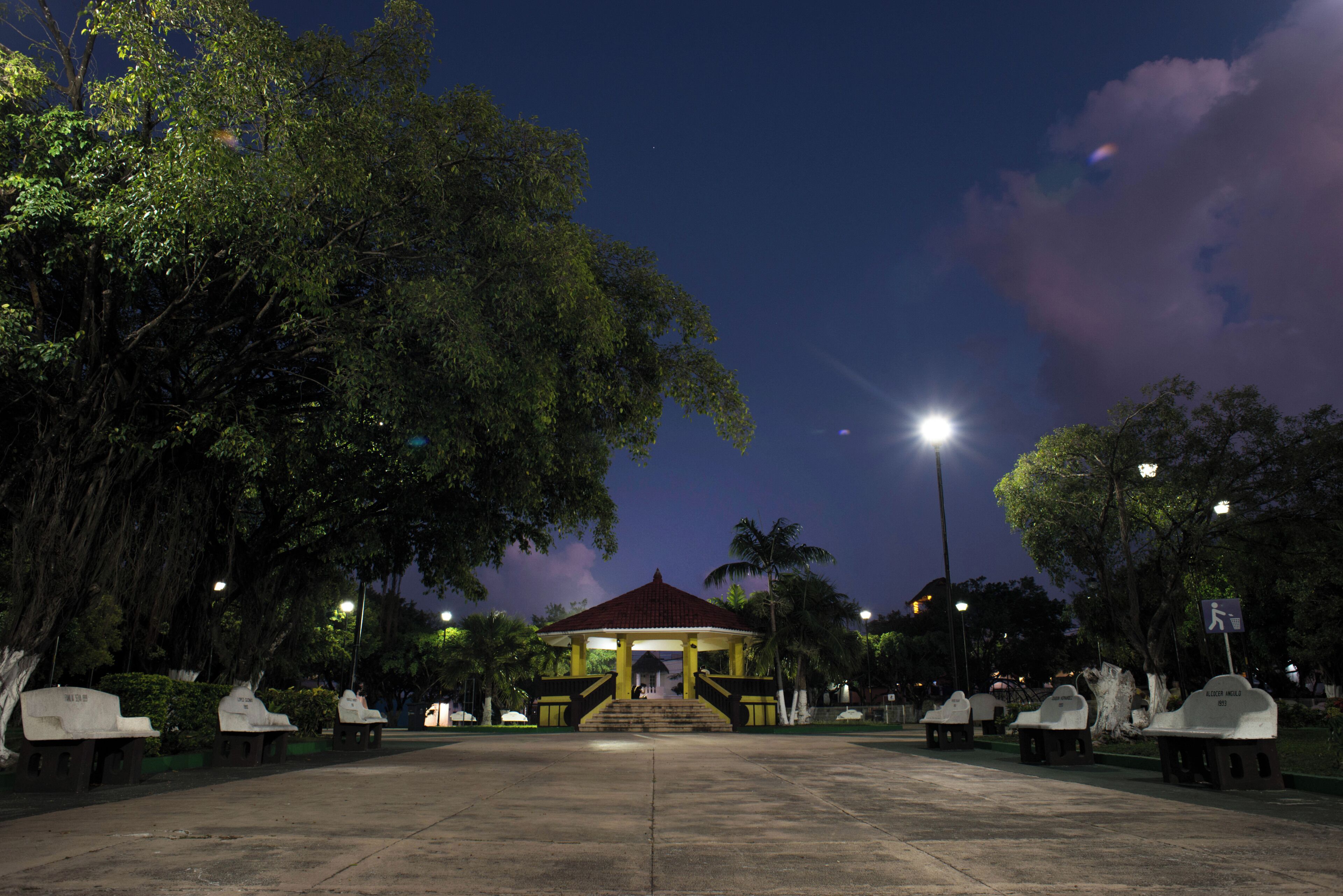 Un gazebo dal tetto rosso, circondato da alberi e panchine, sotto un cielo notturno con nuvole visibili.