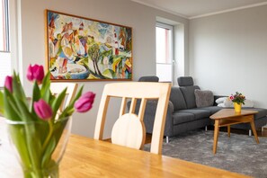 A modern living room with a grey sofa, a wooden chair, a small table, and a colorful abstract painting on the wall. - Dancing lighthouse (Wittdün)