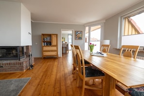 A dining area with a wooden table and chairs, a fireplace, a bookshelf, and a view through a window. - Dancing lighthouse (Wittdün)