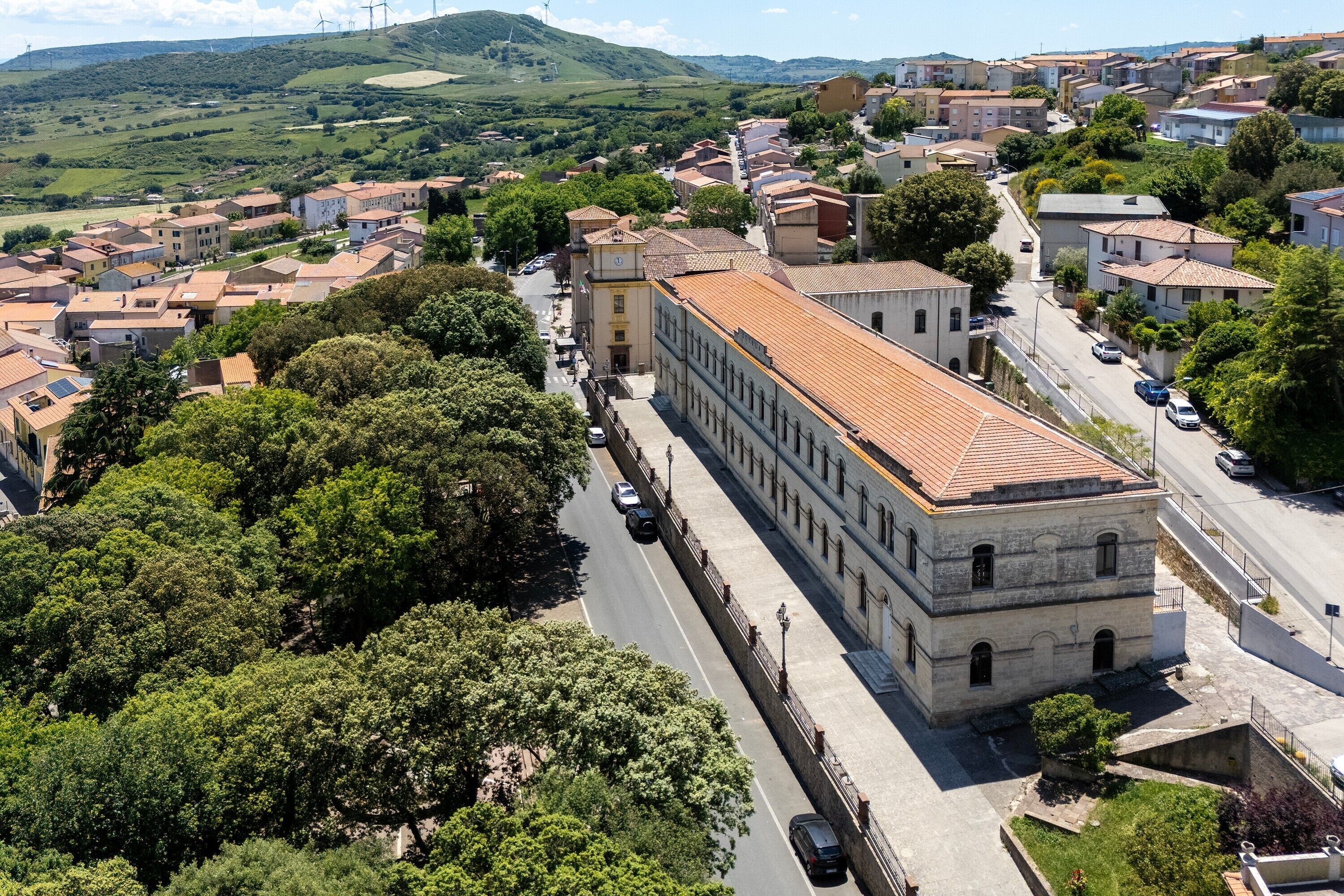 An aerial view of a town with a prominent building with a red-tiled roof, surrounded by greenery and residential buildings.
