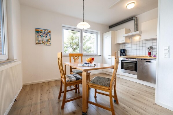 A modern kitchen with white cabinets, a stainless steel oven, and a dishwasher. There is a dining table with two chairs and a pendant light.
