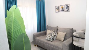 A living room with a grey sofa, a large green plant, blue curtains, and three framed artworks on the wall. - Makasembo Cozy Serenity (Kisumu)