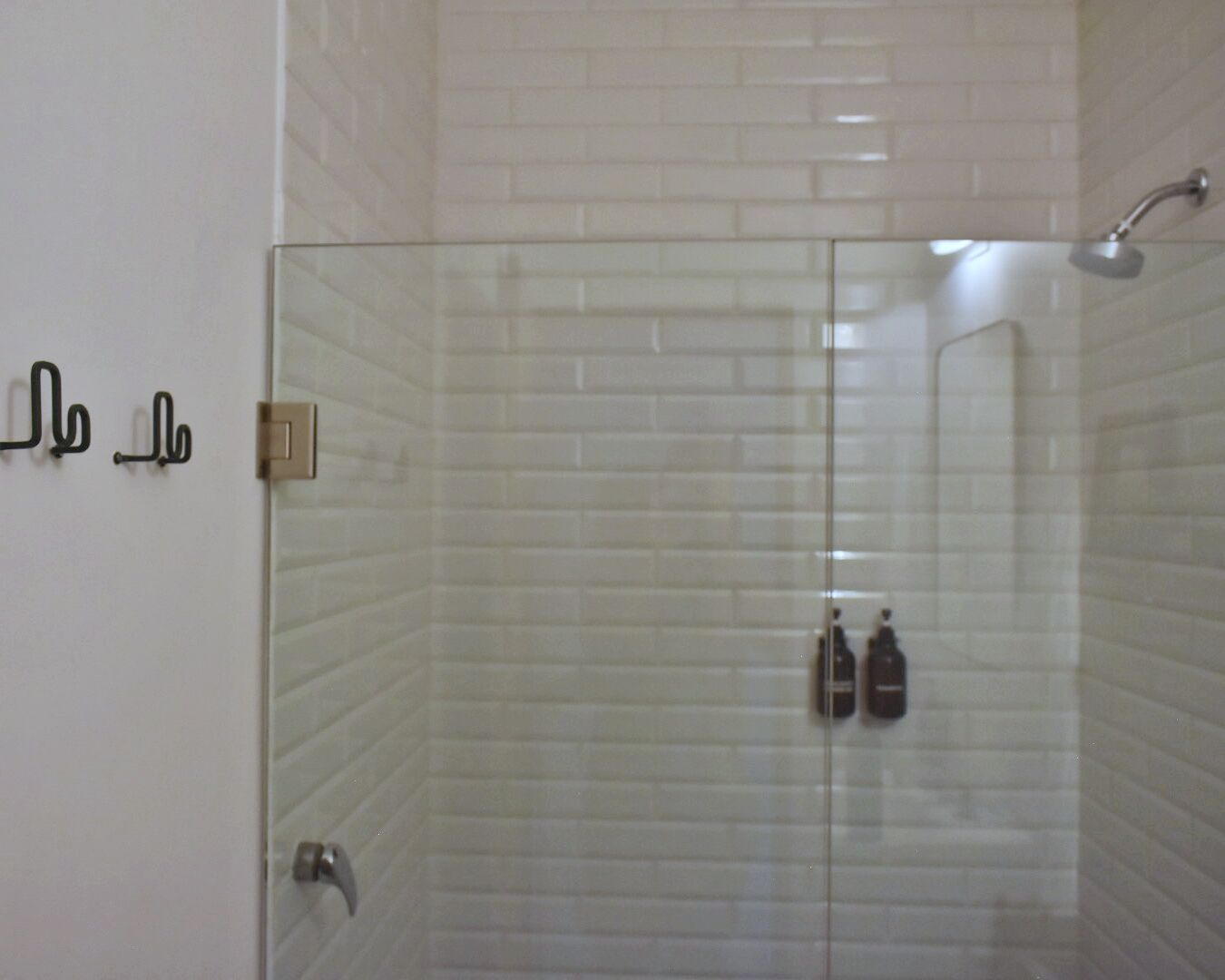 A glass-enclosed shower with white tiles, a showerhead, and two bottles on the wall.