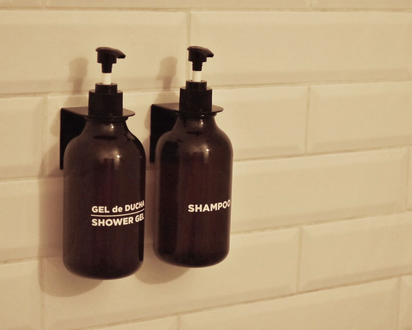 Two dark brown bottles with pumps, labeled 'GEL de DUCHA SHOWER GEL' and 'SHAMPOO,' mounted on a white tiled wall.