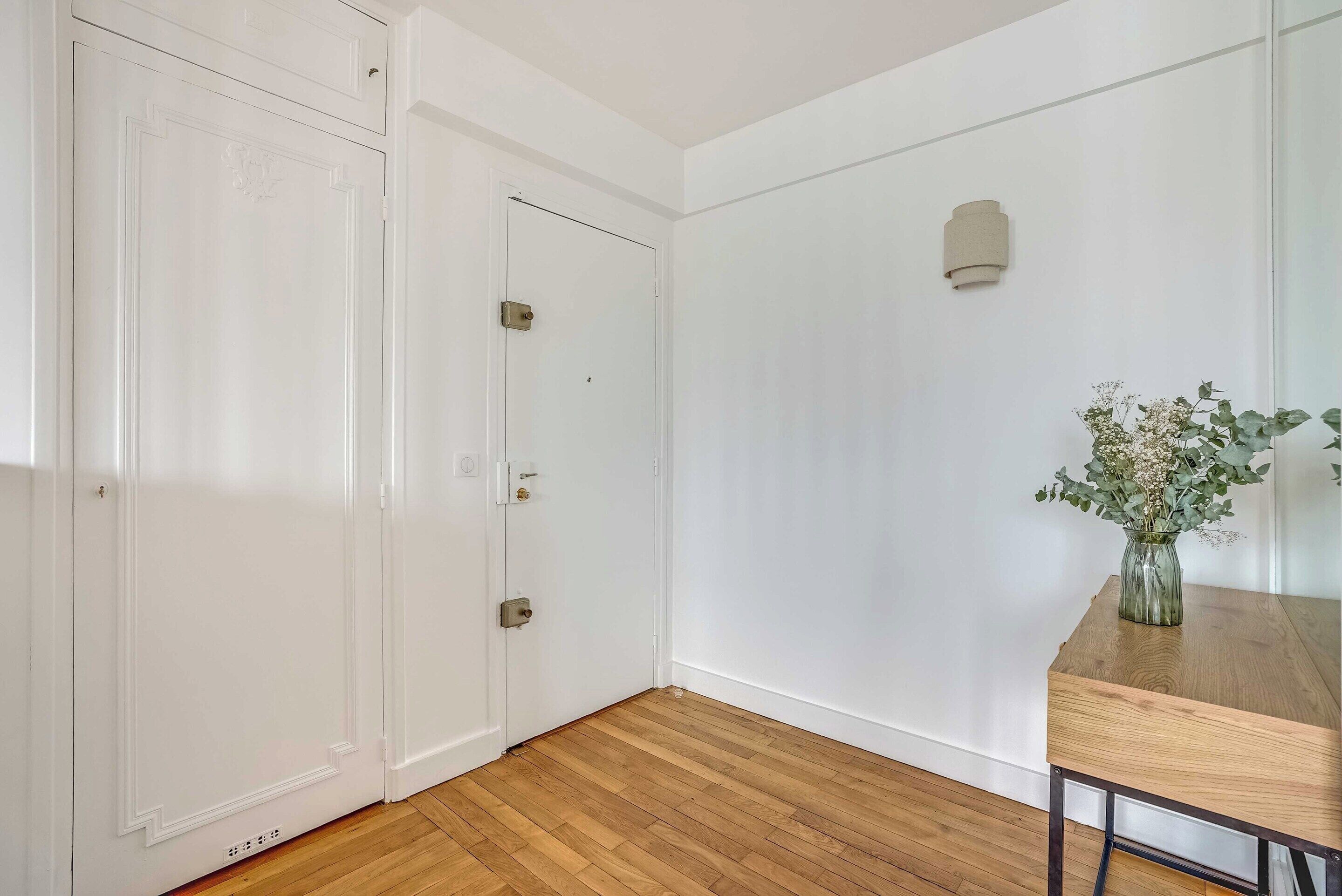A room with a wooden floor, white walls, a wooden table with a vase, and a door with a handle.