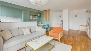 Uma sala de estar com sofá, mesa de centro, espaço de jantar com mesa e cadeiras, e cozinha com armários e luminária pendente.