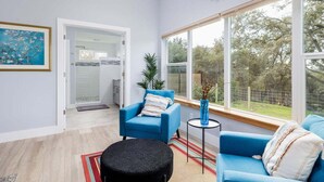 Ein modernes Wohnzimmer mit einem blauen Sofa, einem schwarzen Ottomanen und einem großen Fenster mit Blick auf Bäume.
