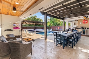A covered outdoor area with a pool, dining table, chairs, wicker furniture, and a kitchen area. - Villa Sunset by Glam Homes (Hialeah)