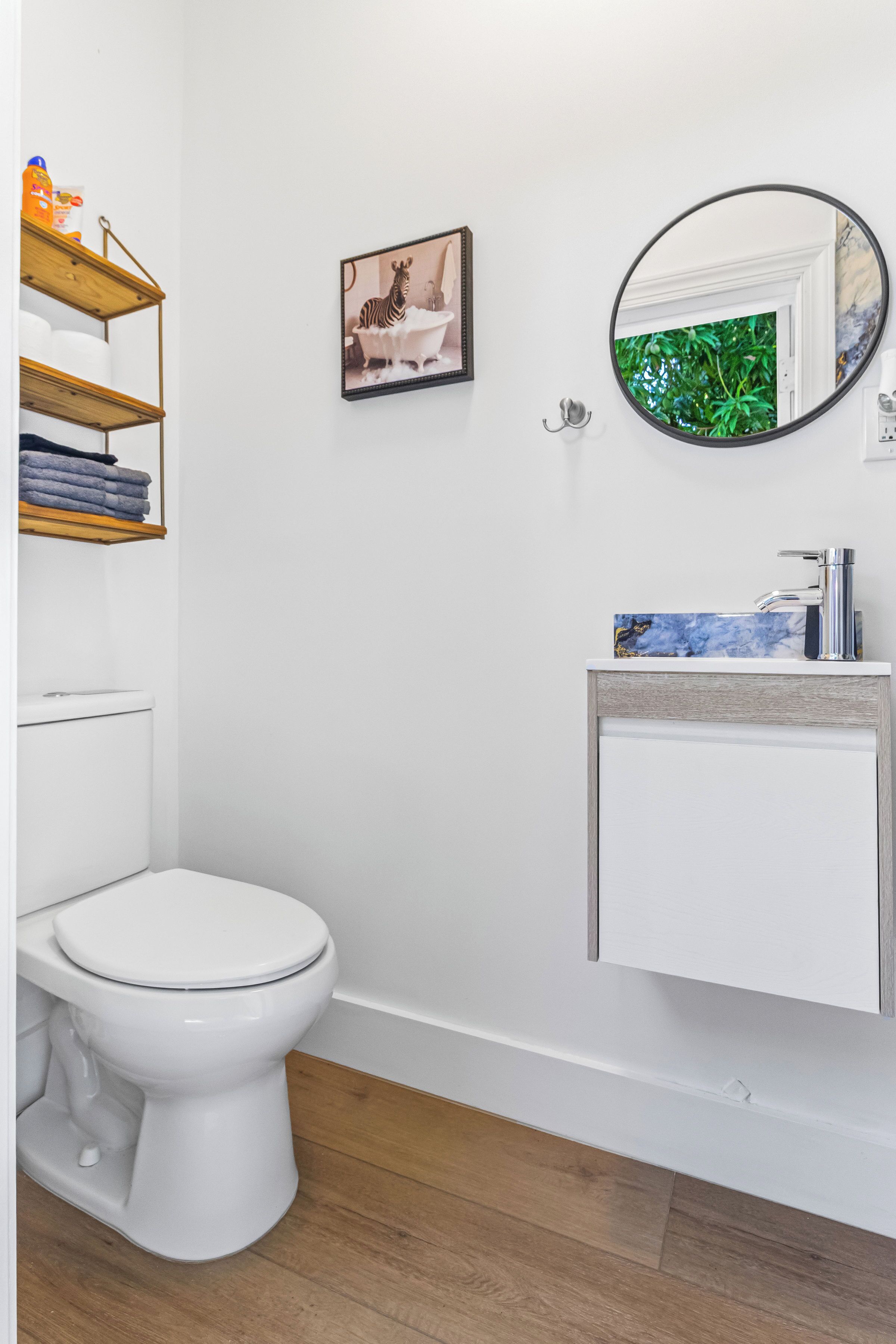 Un baño con un inodoro blanco, estantes de madera, un cuadro enmarco, un espejo redondo y un lavabo con encimera de mármol.