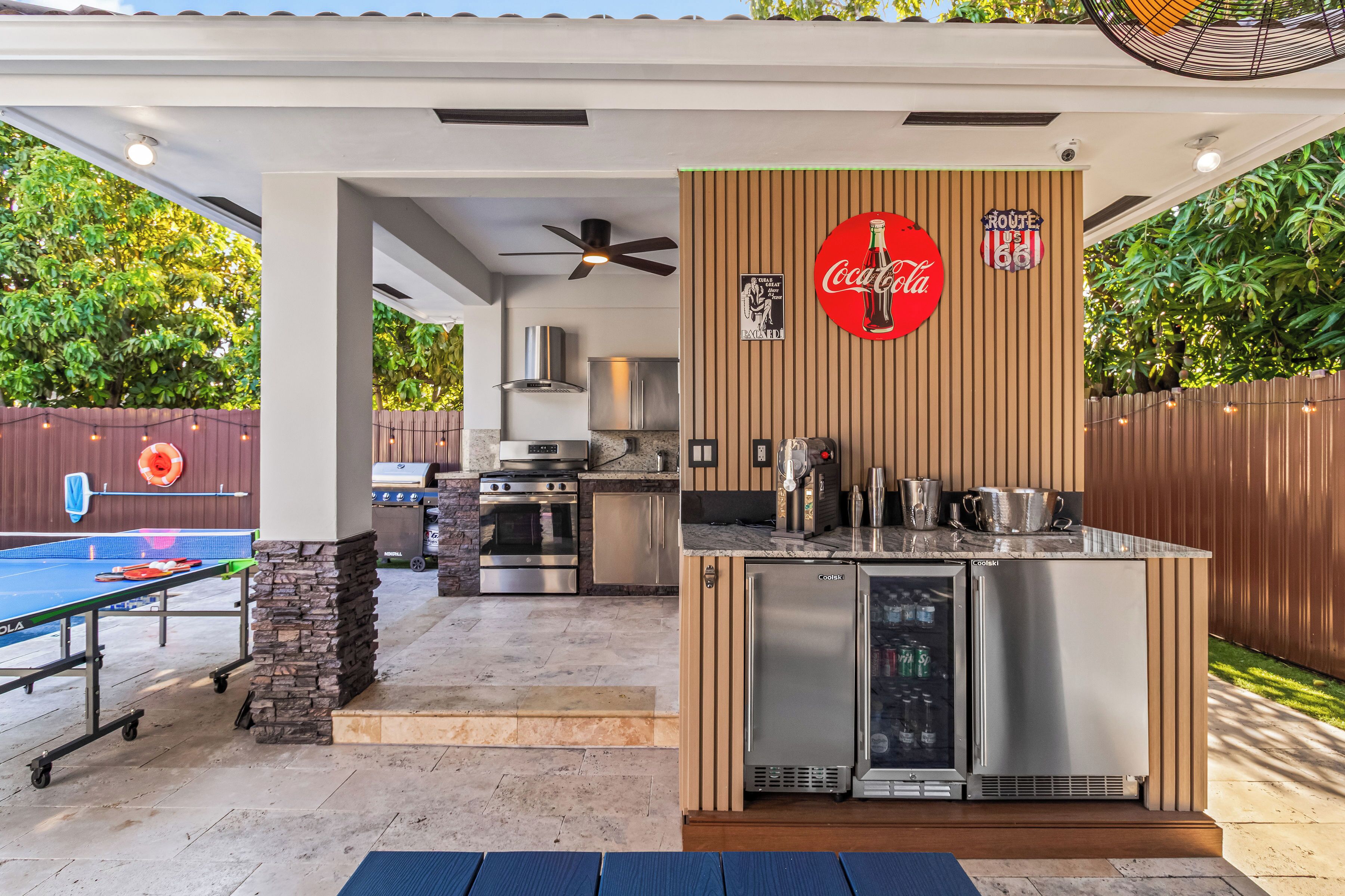 Une cuisine extérieure équipée d’un réfrigérateur, d’une cuisinière et d’un barbecue, ainsi qu’une table de ping-pong et une enseigne Coca-Cola.