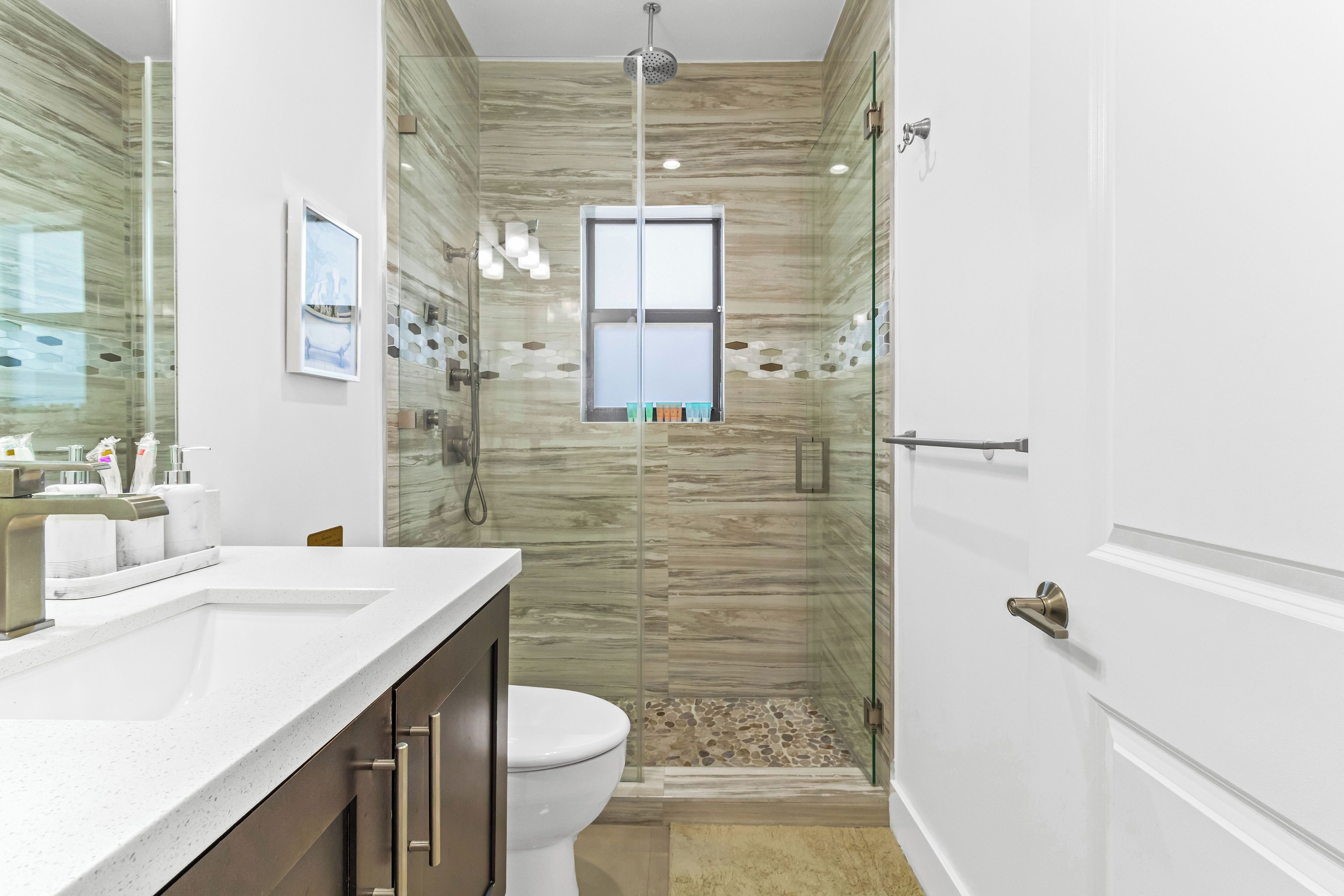 A bathroom with a glass shower enclosure, a toilet, a sink with a marble countertop, and a window with a view.