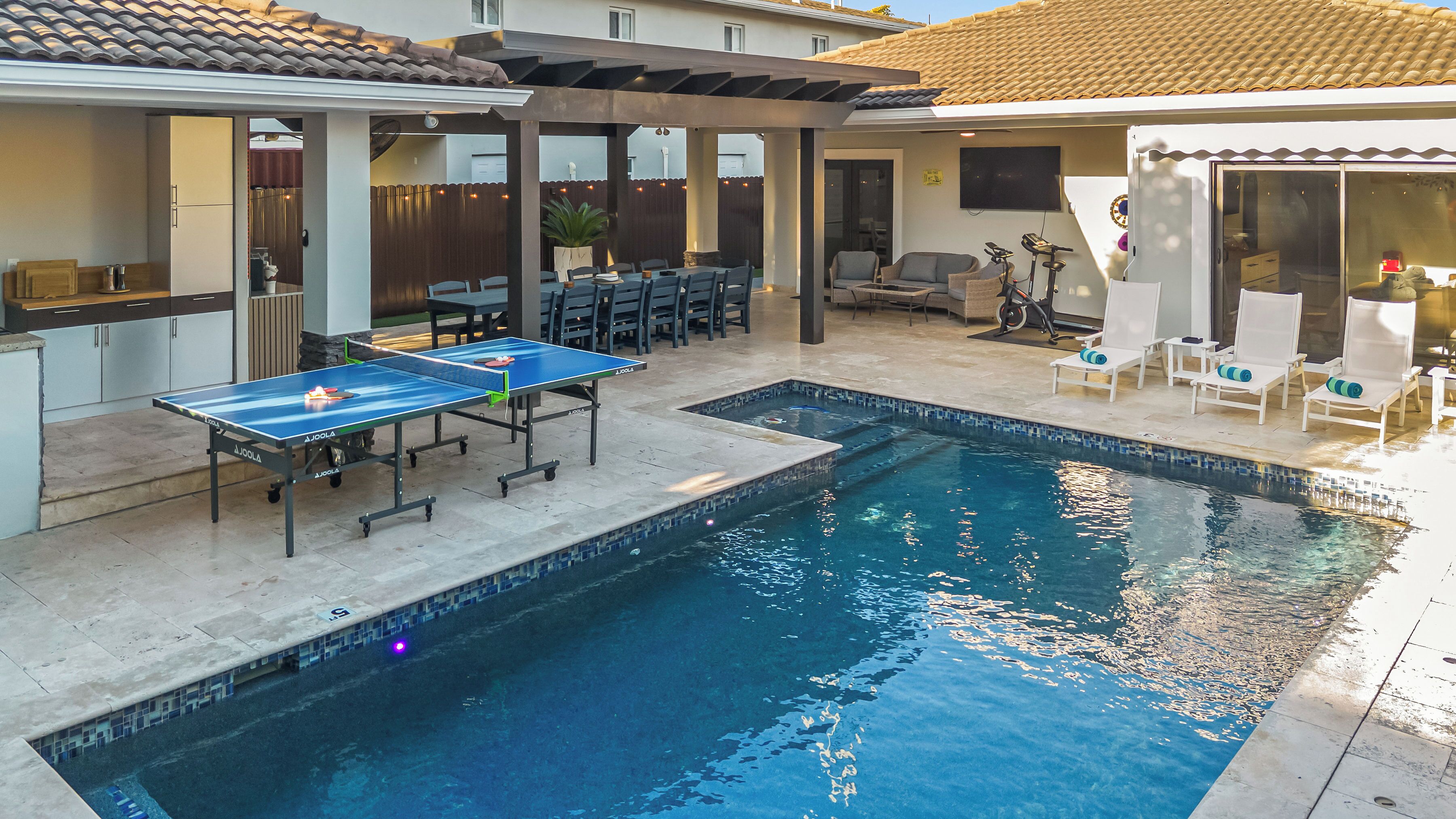 A swimming pool with a ping pong table, outdoor seating, and a covered patio area.