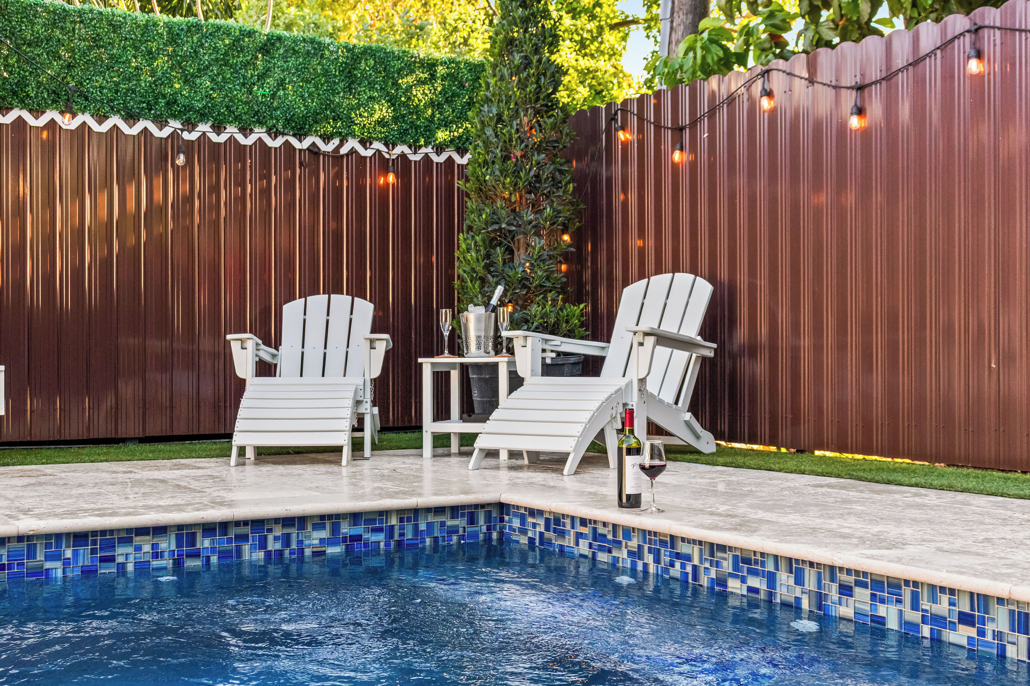 Una piscina con bordes de azulejos, rodeada por una valla de madera, y dos sillones blancos con una mesa y una botella de vino.