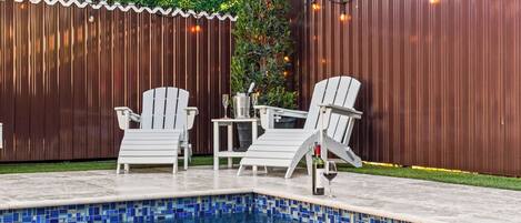 Una piscina con bordes de azulejos, rodeada por una valla de madera, y dos sillones blancos con una mesa y una botella de vino.