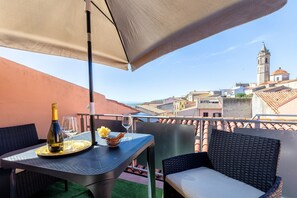 Un balcón con una mesa, una botella y copas, con vistas a un paisaje urbano con edificios y una torre.