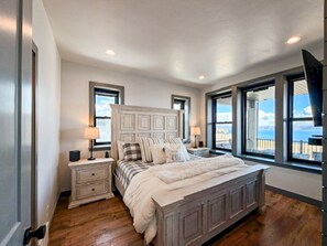 A bedroom with a large bed, two bedside tables, a headboard, and a view through large windows. - Bear Lake Modern Family Retreat (Garden City)