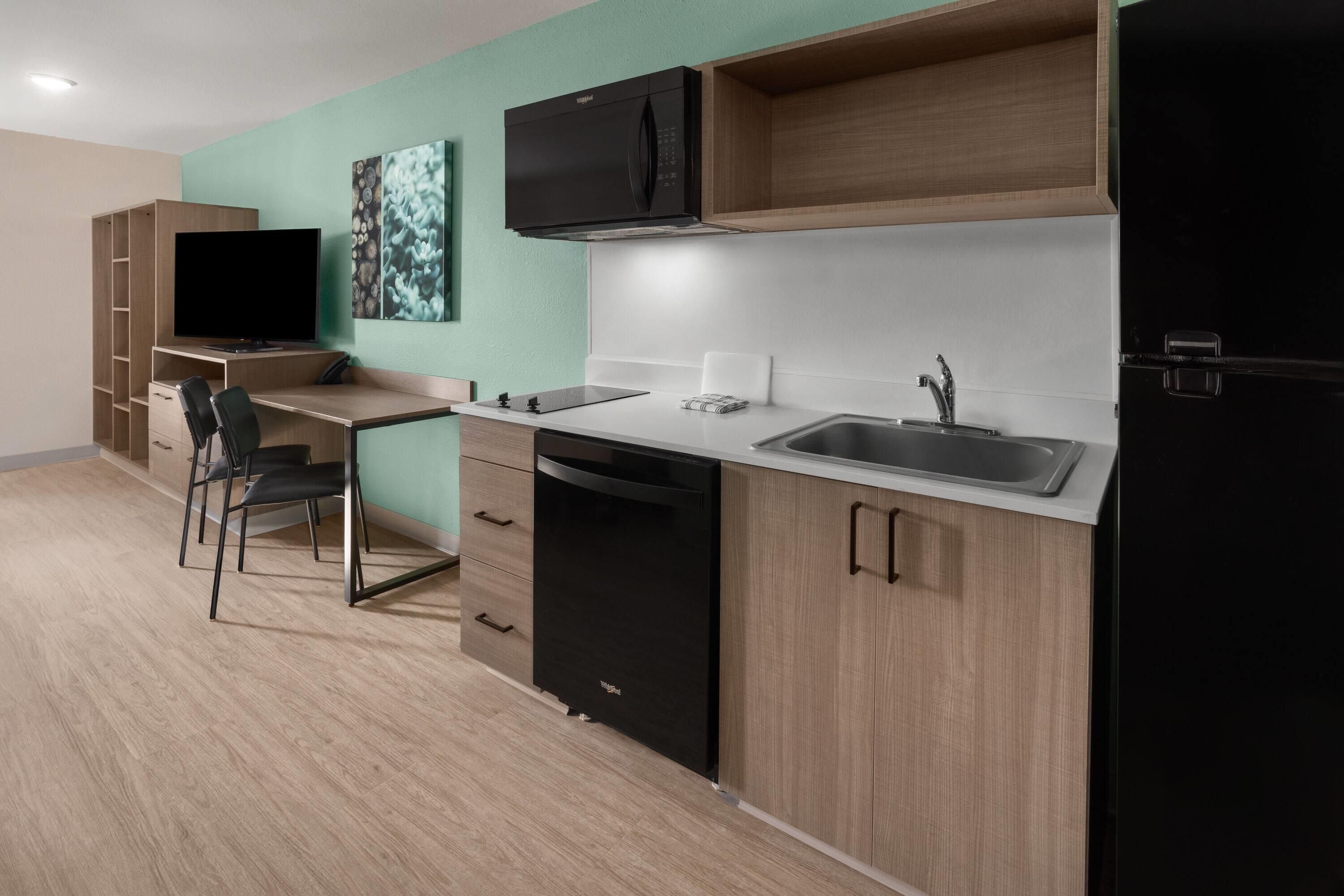 A modern kitchenette with a built-in microwave, stove, and sink. There is a small dining area with a chair and a TV.