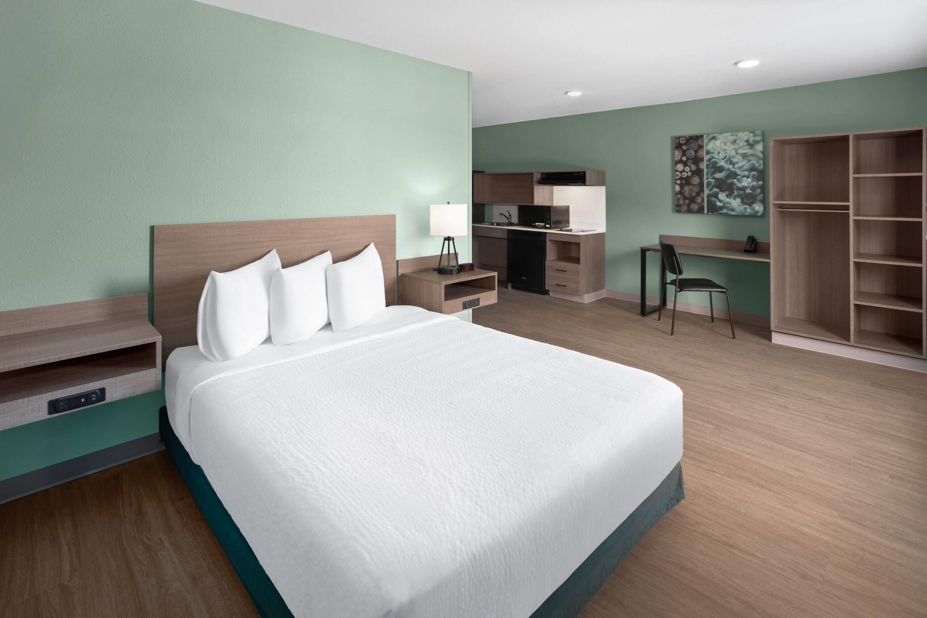 A hotel room with a large bed, a desk, a kitchenette, and a wardrobe.