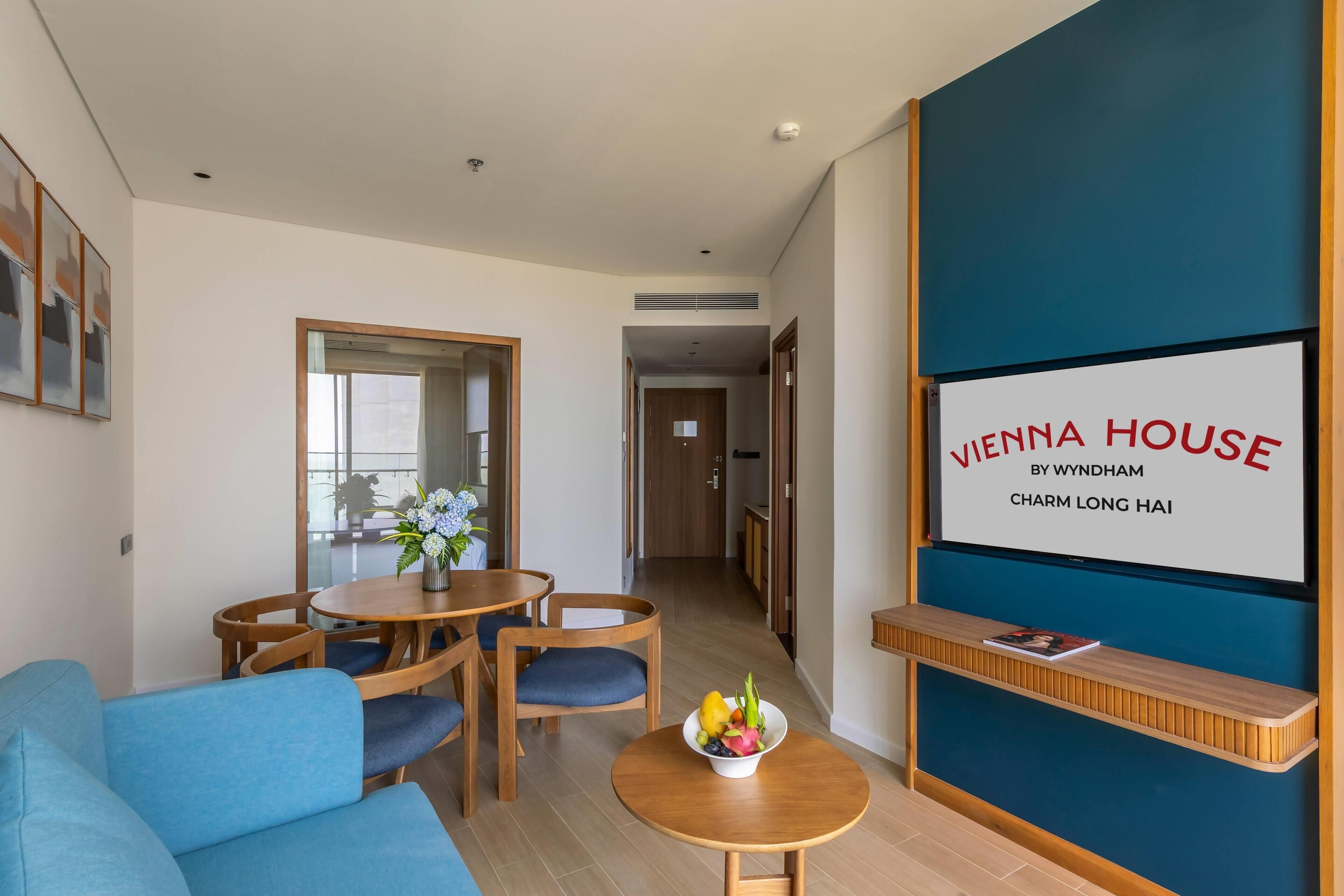 Moderni hotellihuone, jossa on sininen tehosteseinä, ruokailutila puupöydällä ja -tuoleilla, pienellä pöydällä oleva hedelmäkulho sekä kyltti, jossa lukee: »Vienna House by Wyndham Charm Long Hai».