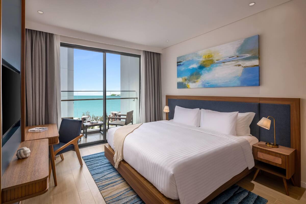 A hotel room with a large bed, a desk, a chair, a nightstand, and a painting on the wall.