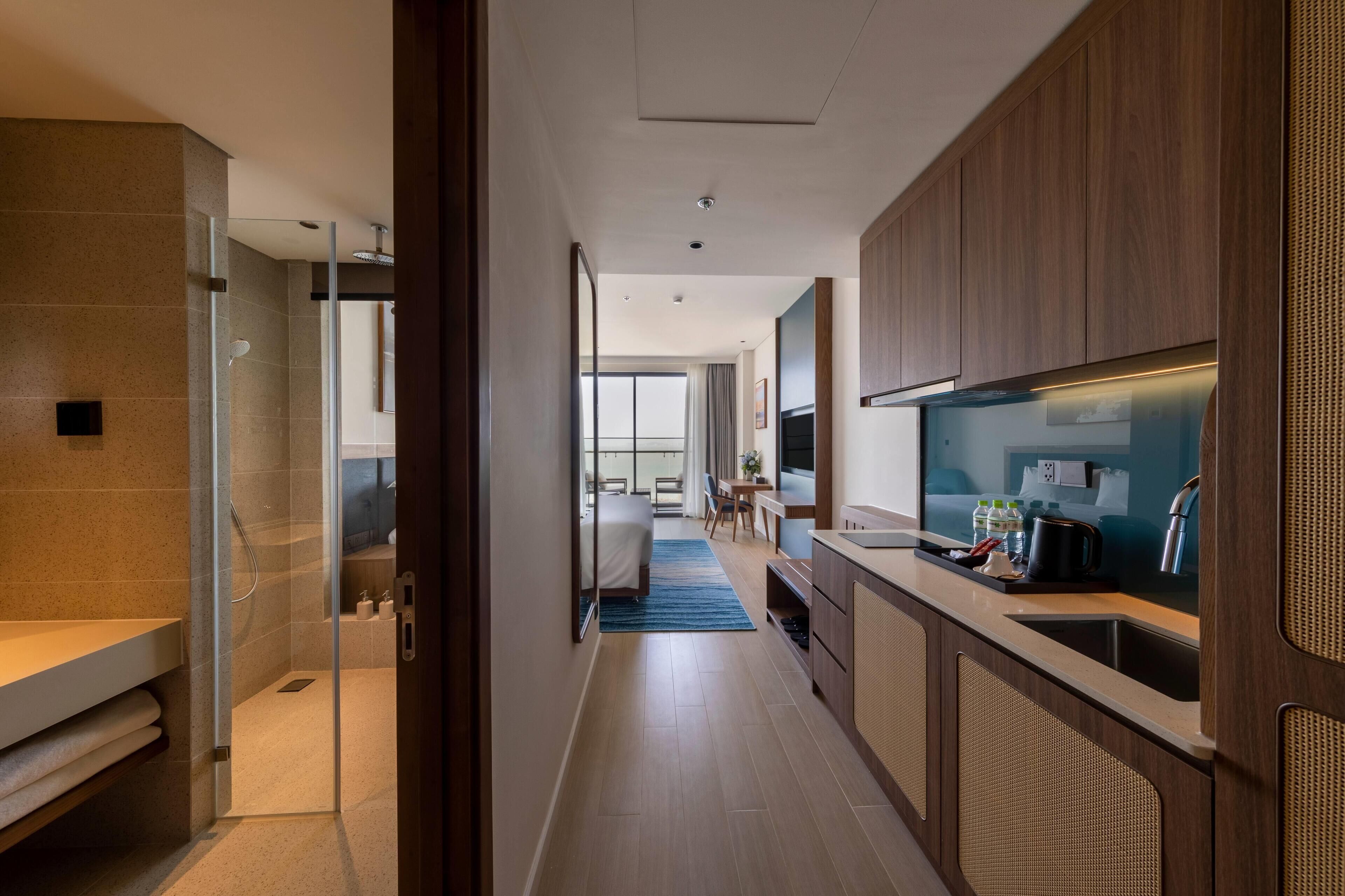 Quarto de hotel moderno com chuveiro, cama, área de refeições e cozinha compacta.