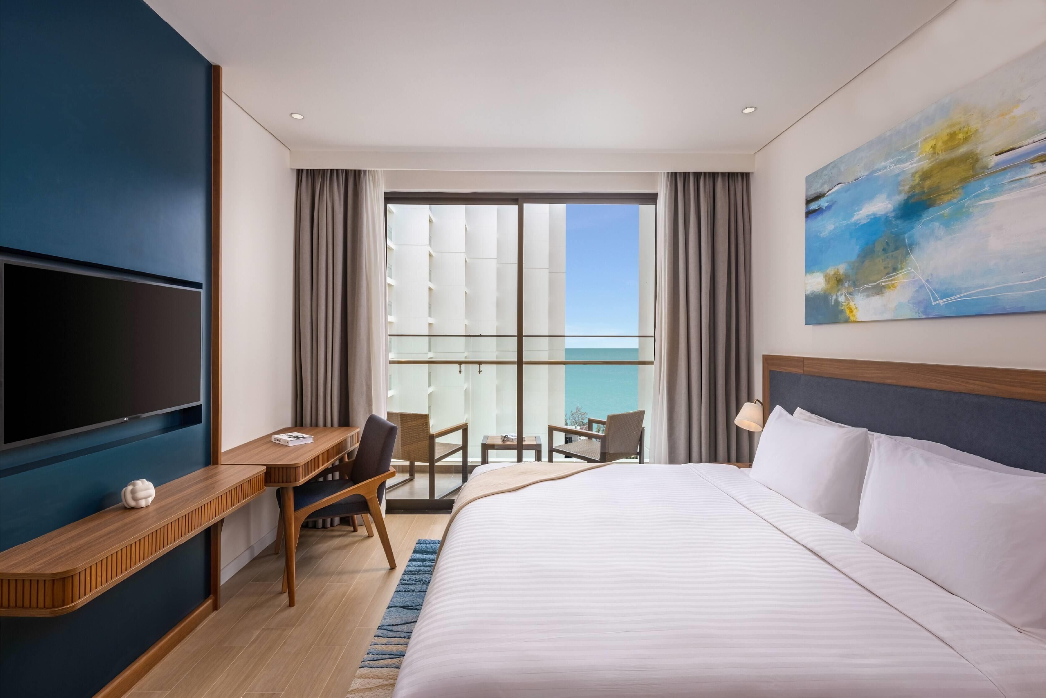 A hotel room with a large bed, a TV, a desk with a chair, a balcony with a view, and a painting on the wall.