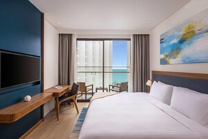 A hotel room with a large bed, a TV, a desk with a chair, a balcony with a view, and a painting on the wall.