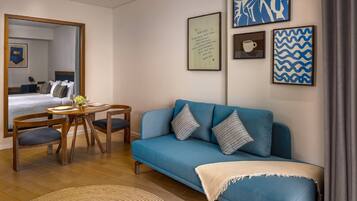 A modern hotel room with a blue sofa, wooden dining table, and chairs, a bed with a view, and artwork on the walls.