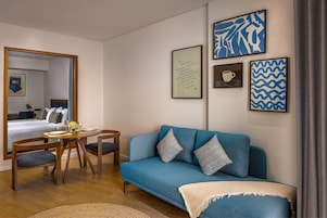 A modern hotel room with a blue sofa, wooden dining table, and chairs, a bed with a view, and artwork on the walls.