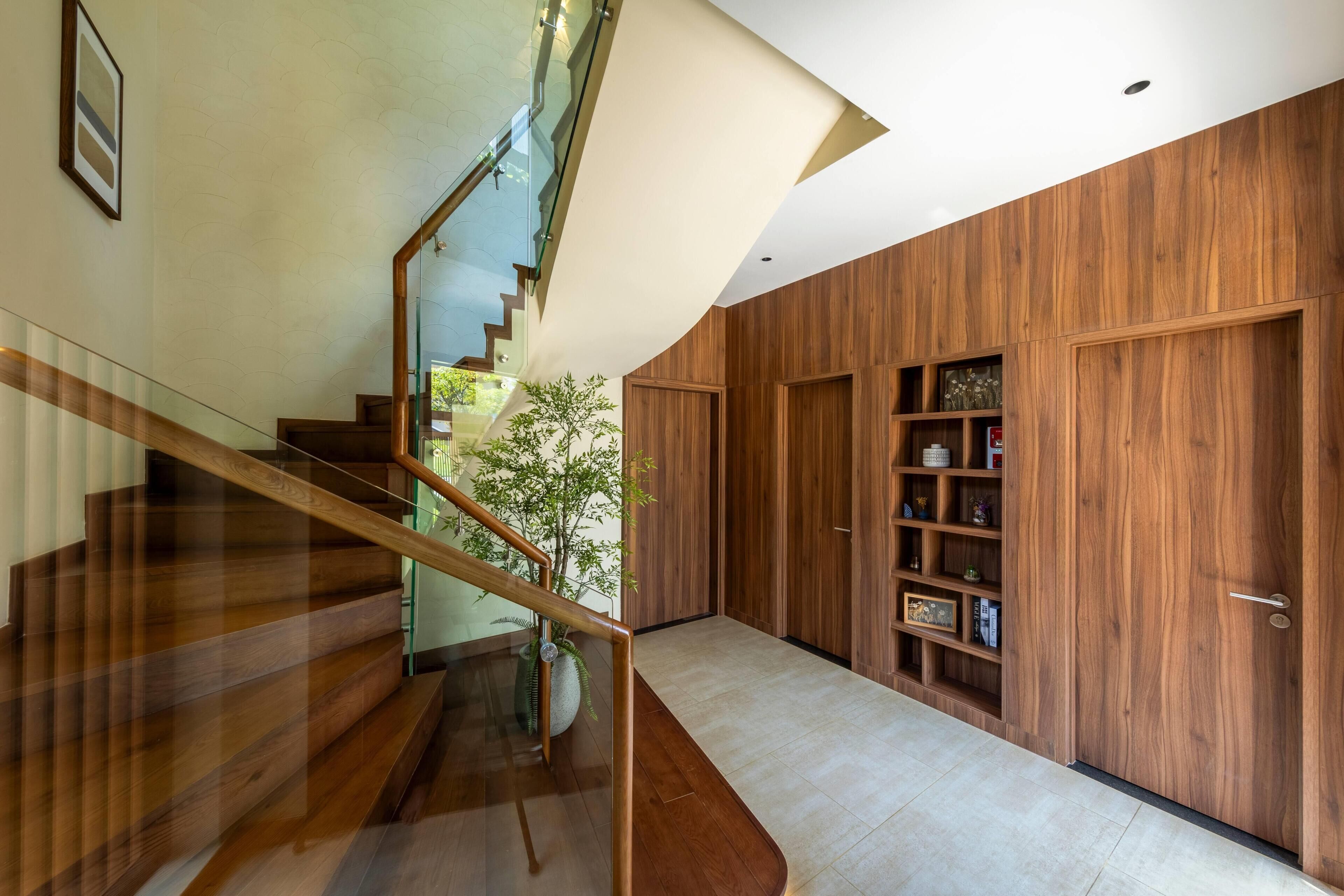 Un interior moderno con una escalera de madera, barandilla de cristal y una puerta de madera con estante incorporado.