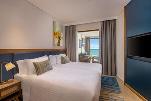 Ett hotellrum med en stor säng, en TV, ett skrivbord och utsikt över havet.
