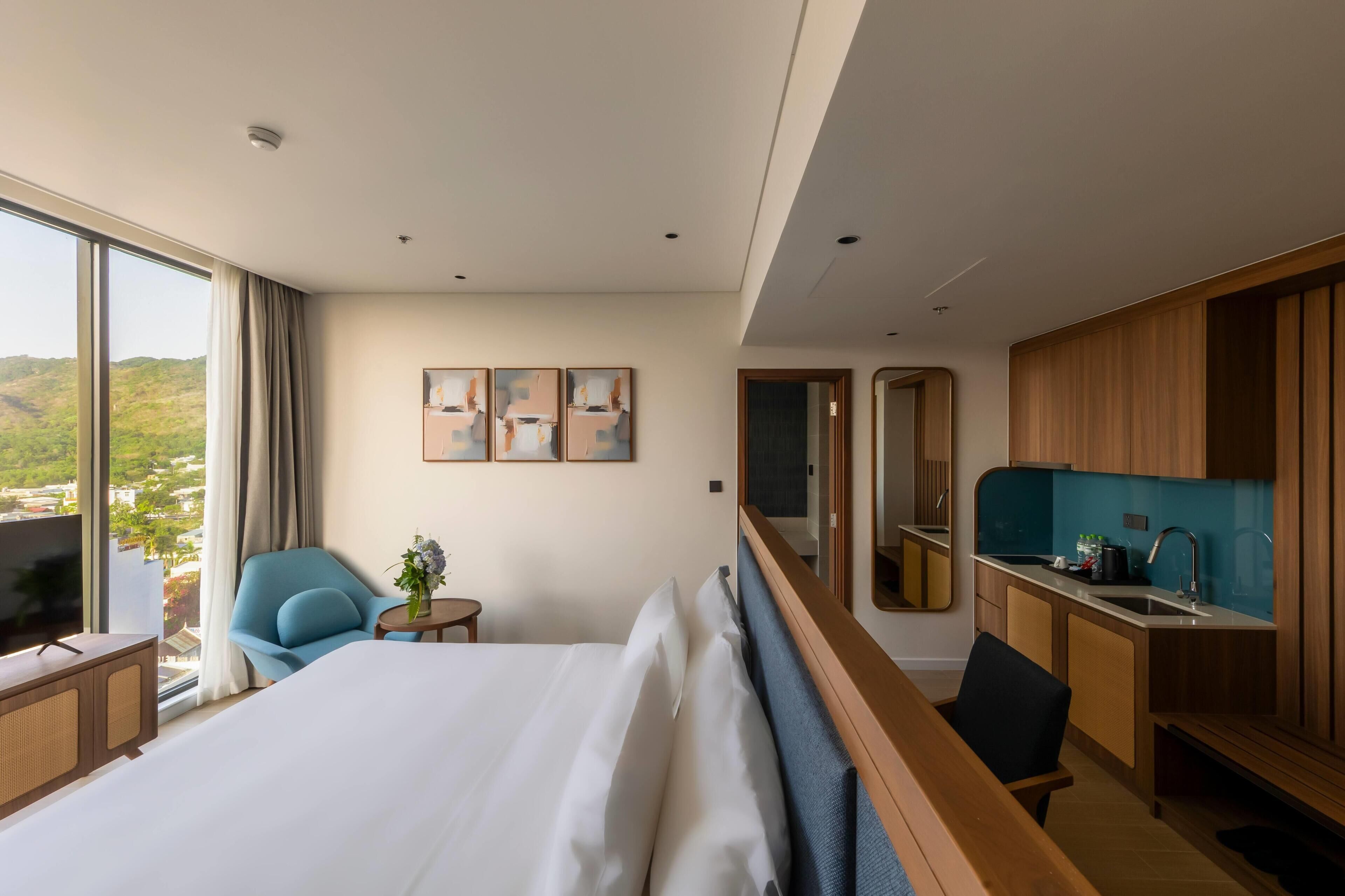 A hotel room with a large bed, a TV, a desk, a chair, a small table with a plant, and a view of the outdoors.