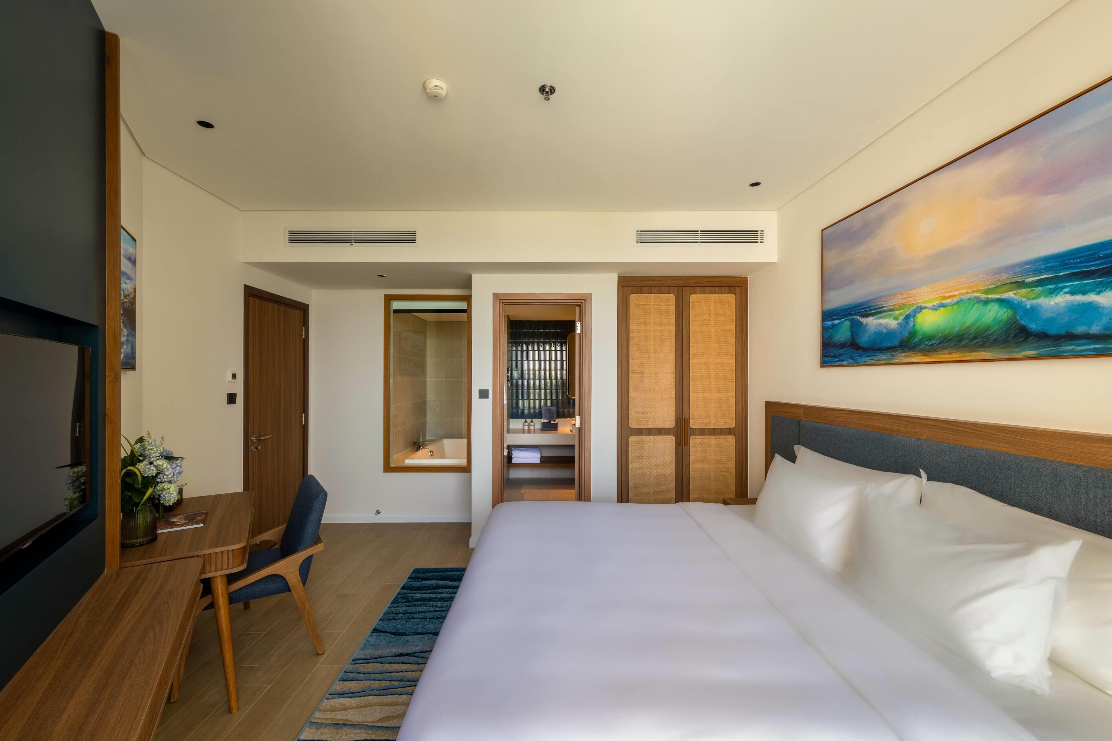A hotel room with a bed, a desk, a chair, a TV, a painting, and a bathroom visible through an open door.