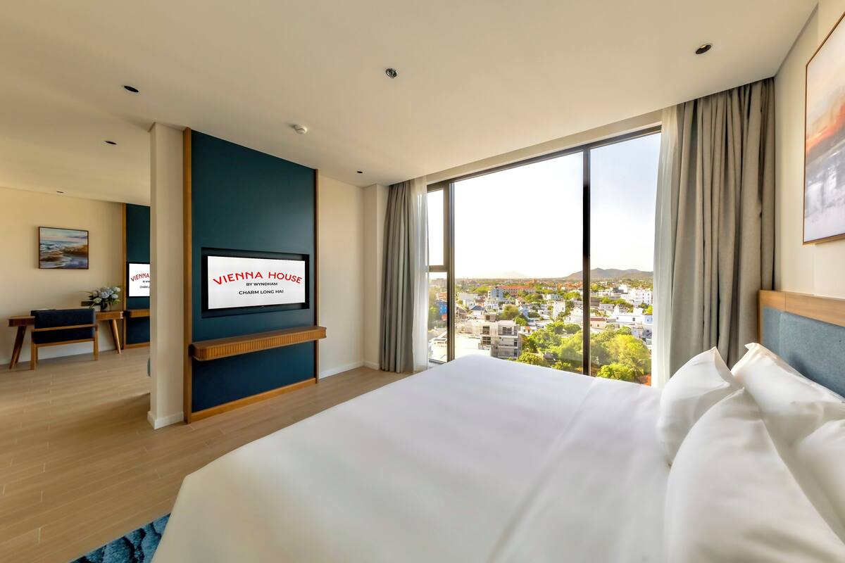 A hotel room with a large bed, a television displaying 'VIENNA HOUSE BY WYNDHAM CHARM LONG HALL,' a desk with a chair, and a large window offering a city view.