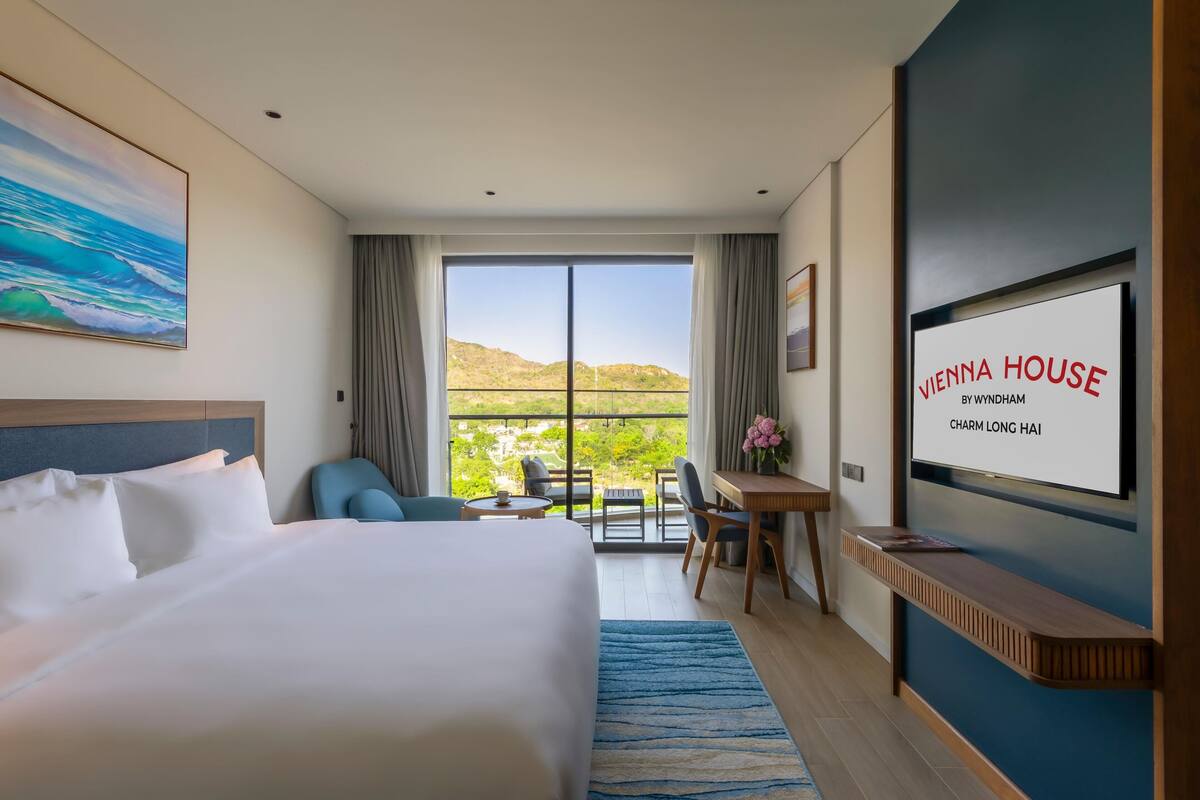 A hotel room with a large bed, a desk, a chair, a TV, and a balcony with a view.