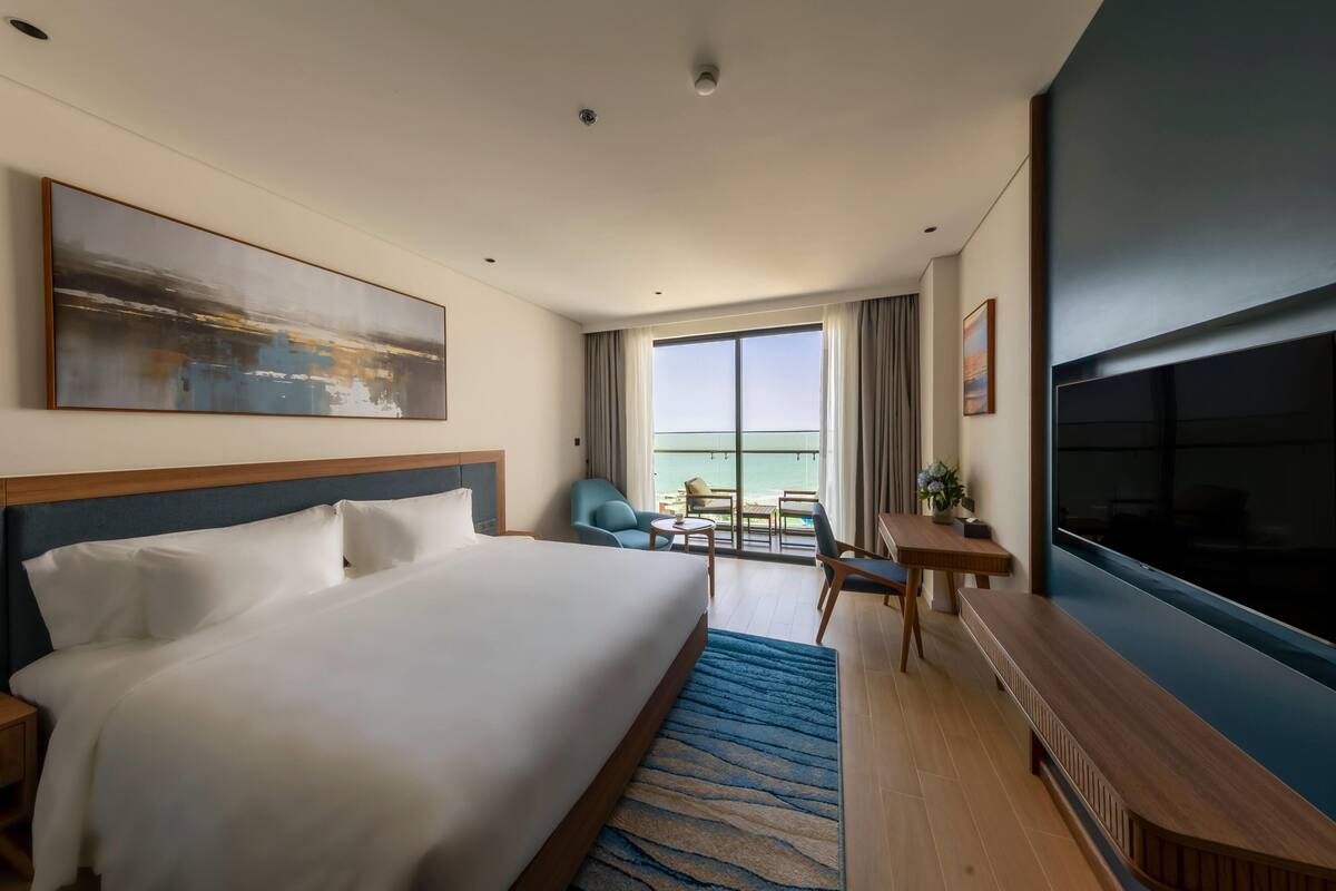 A hotel room with a large bed, a desk with a chair, a TV, and a balcony with a view.