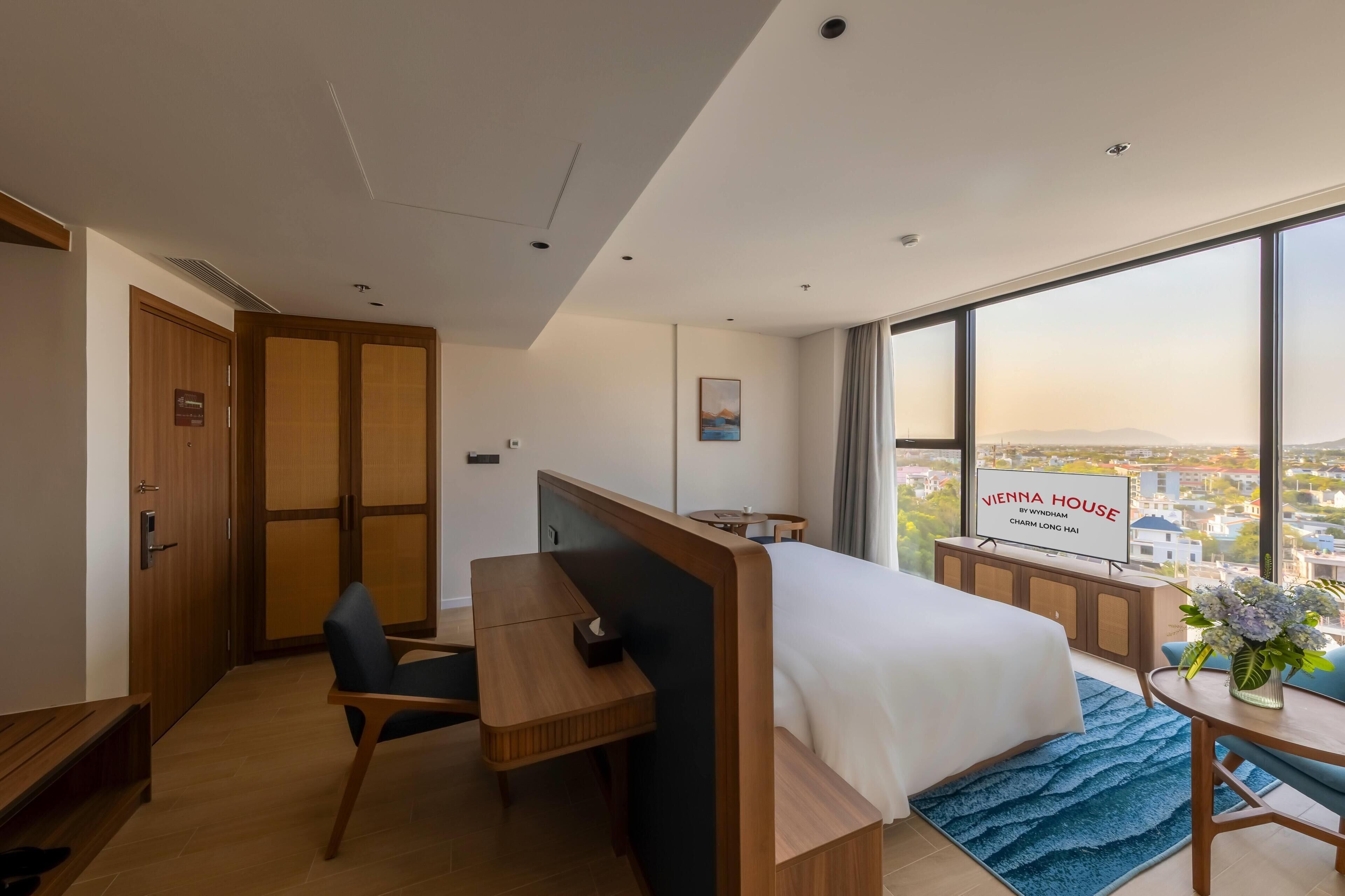 Ein Hotelzimmer mit Bett, Schreibtisch, Stuhl, Fernseher und einem großen Fenster mit Blick auf die Stadt.