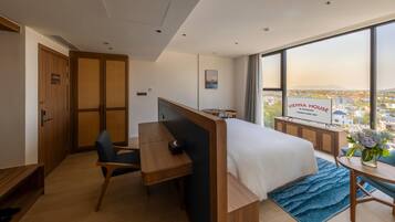 Ein Hotelzimmer mit Bett, Schreibtisch, Stuhl, Fernseher und einem großen Fenster mit Blick auf die Stadt.