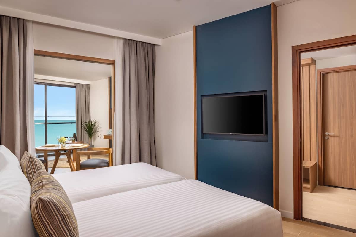 A hotel room with two beds, a TV, a balcony with a table and chairs, and a view of the sea.