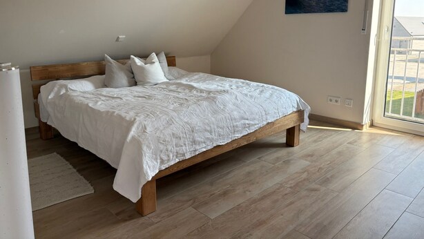Una camera da letto con un letto di legno, biancheria bianca, vista attraverso una grande finestra e un quadro appeso al muro.