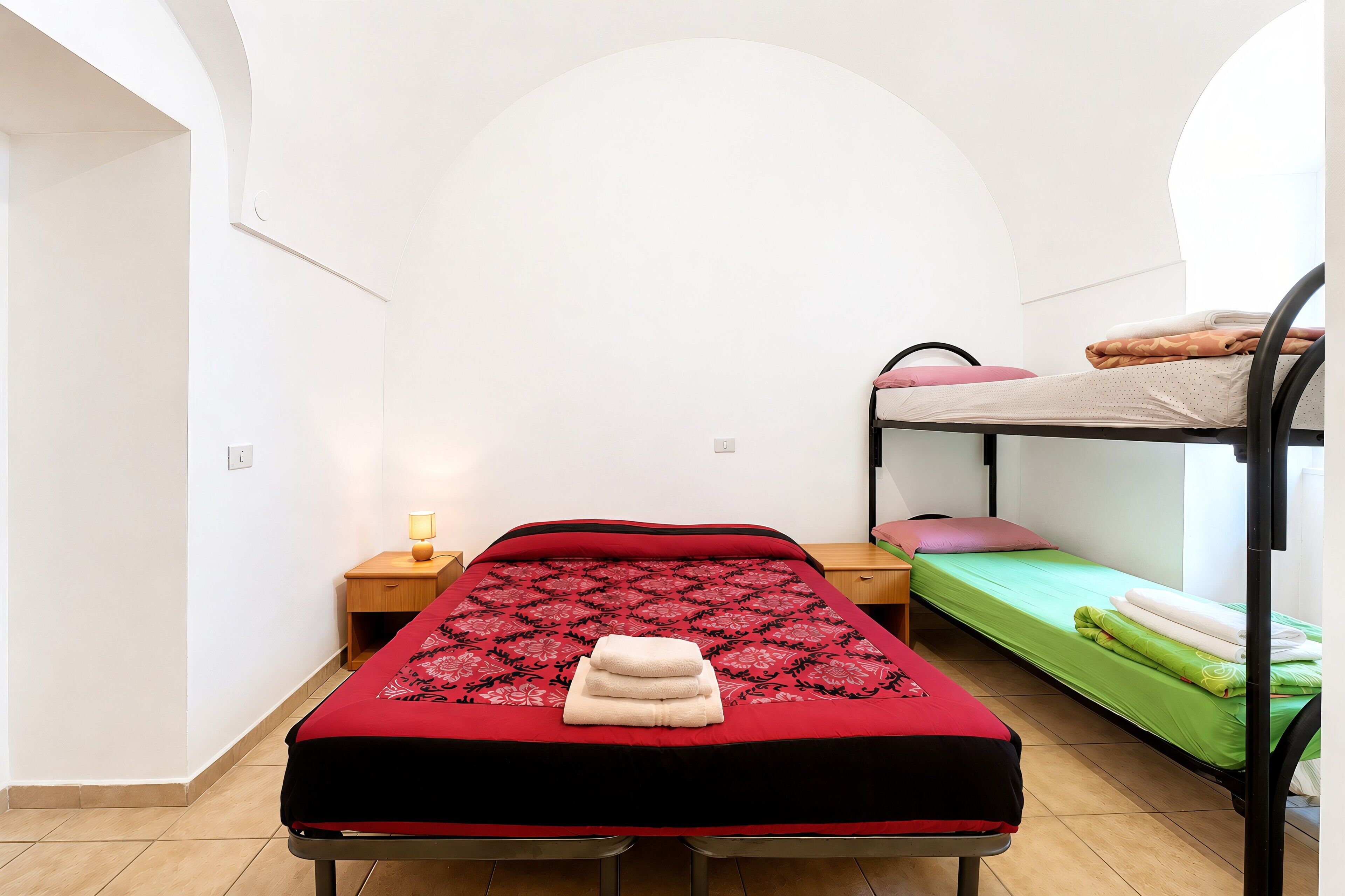 A single bed with a red and black bedspread, a bedside table with a lamp, and a bunk bed to the right.